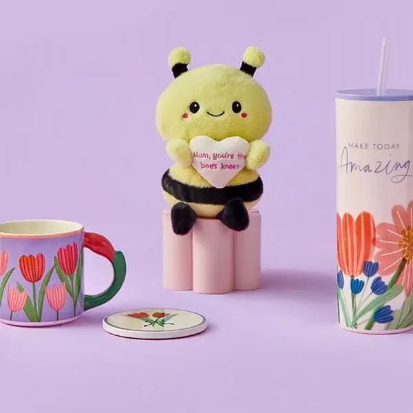 A cute bee plush holding a heart sits between a tulip‑patterned mug, a floral coaster, and a tall floral tumbler with a straw, all arranged on a soft pastel purple background.
